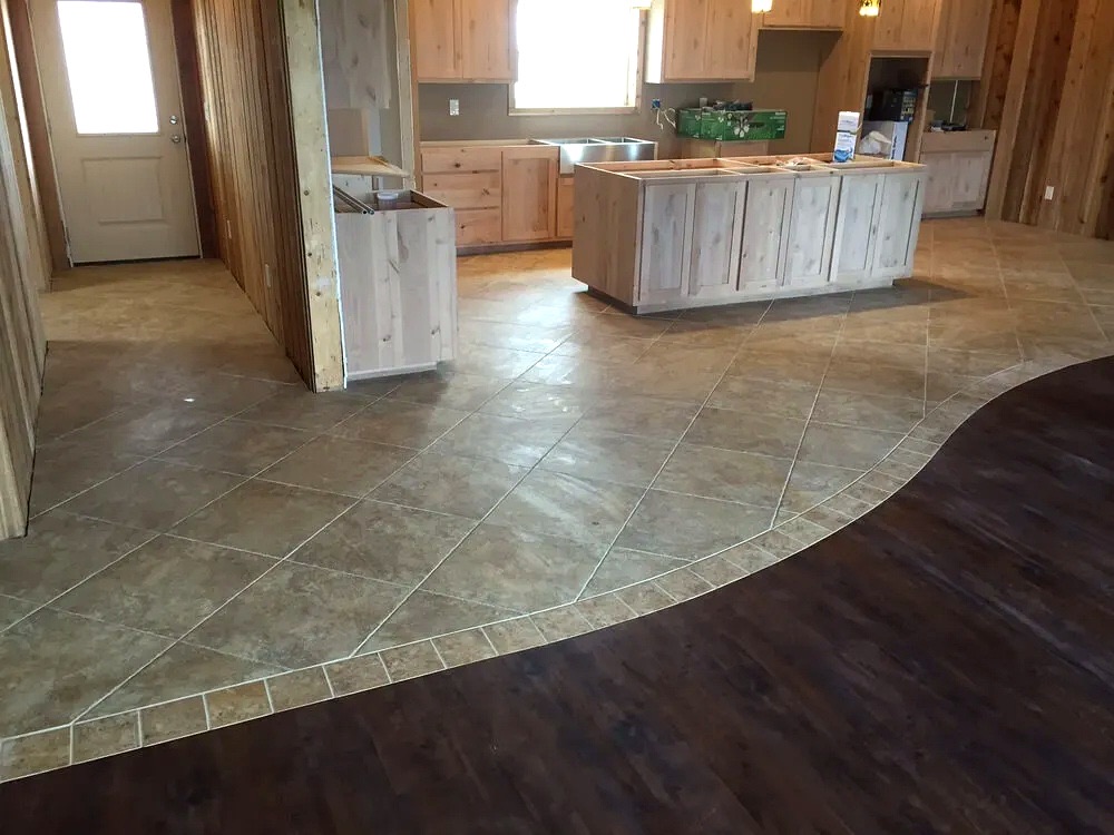 Open kitchen under construction with diagonal tile flooring that curves into dark plank flooring, separated by a tile border